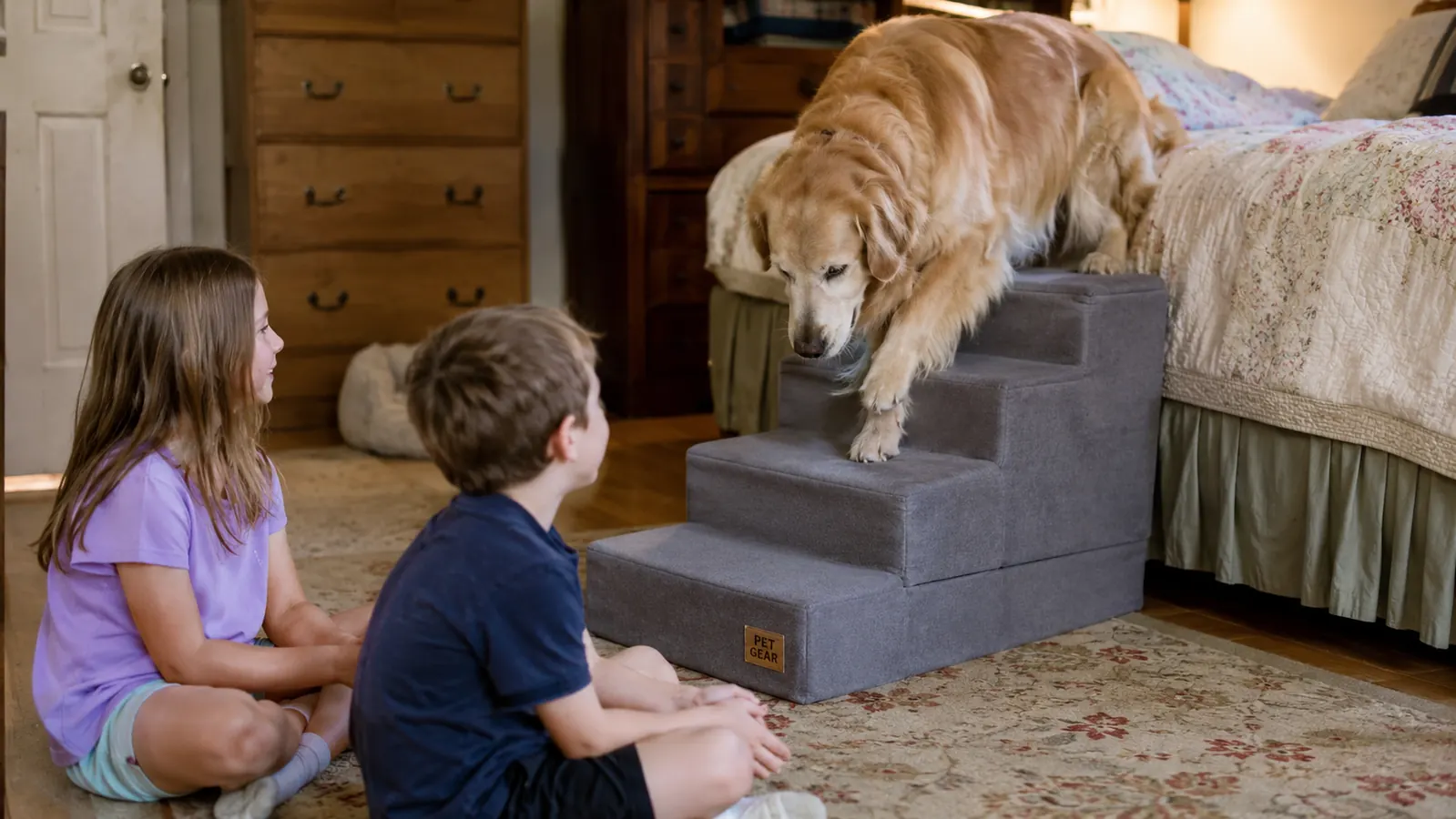 Grandkids sitting on a bedroom floor watching a golden retriever walk down a set of grey dog stairs