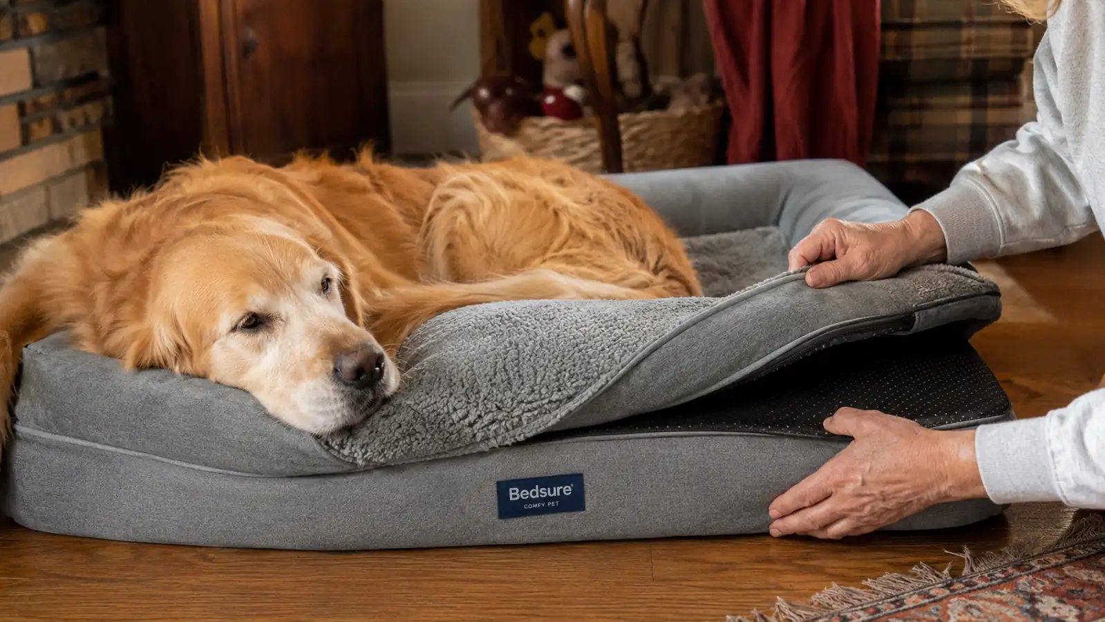 Hands smoothing the removable cover onto a gray orthopedic dog bed with bolster sides on a hardwood floor