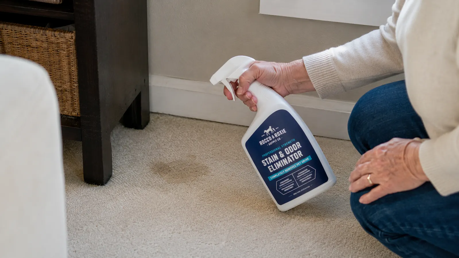 An older woman's hand spraying a clear enzyme cleaner from a labeled bottle onto a beige carpet stain
